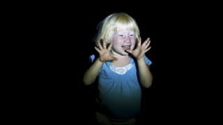 Ein hellblondes Kind steht im Dunkeln und wird angeleuchtet, es hält die Hände mit gespreizten Fingern in die Höhe.