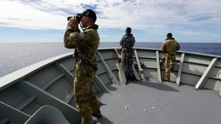 Drei Soldaten blicken vom Bug eines Militärschiffes mit Feldstechern hinaus aufs Meer.