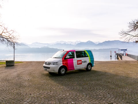 Der SRF Kids Bus steht vor einem See – im Hintergrund sieht man eine Berglandschaft.