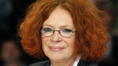 Eine Frau mit roten Locken (auf Kinnhöhe geschnitten), Brille und dunkel geschminkte Augen.