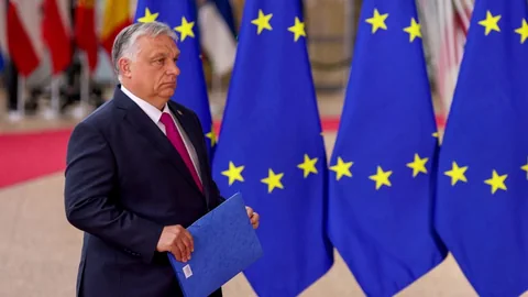 Viktor Orbán geht an EU-Fahnen vorbei und trägt dabei eine blaue Mappe mit sich.