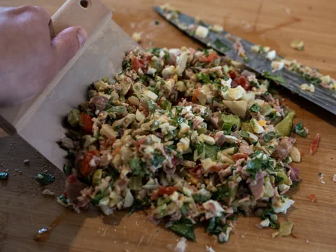 Hand hält Holzspachtel mit gehacktem Gemüse und Wurst auf Schneidebrett.