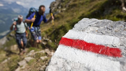 Zwei Wanderer laufen den Berg hoch. Im Vordergrund ist eine weiss-rot-weisse Wanderwegmarkierung auf einem Stein.