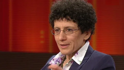 mittelalter Frau mit dunklen, kurzen Locken und ovaler Brille, trägt Blazer und blickt leicht nach rechts.