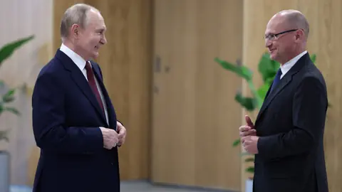 Putin und Kirijenko im Anzug im Gespräch in einem Büro mit Pflanzen.