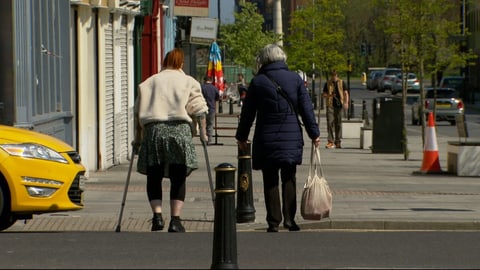 Zwei ältere Personen, eine mit Einkaufssäcken, die andere an Krücken, über die Strasse.