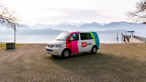 Der SRF Kids Bus steht vor einem See, im Hintergrund sieht man eine Berglandschaft.