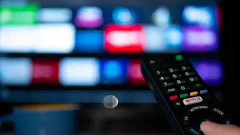 Eine Hand hält eine TV-Fernbedienung, im Hintergrund sind verschwommen bunte Apps auf einem TV-Bildschirm.