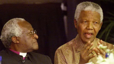 Ein Dunkelhäutiger Mann, Tutu,  schaut nach links zu seinem Freund, Nelson Mandela. Dieser hat den Mund offen.