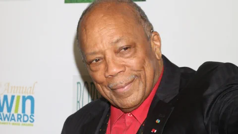 Quincy Jones in einem schwarzen Anzug mit rotem Hemd vor einer weissen Fotowand.