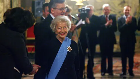 alte Frau mit blauem Band umgelegt, darauf goldene Brosche. Andere Frau schüttelt ihr die Hand, dahinter Leute.