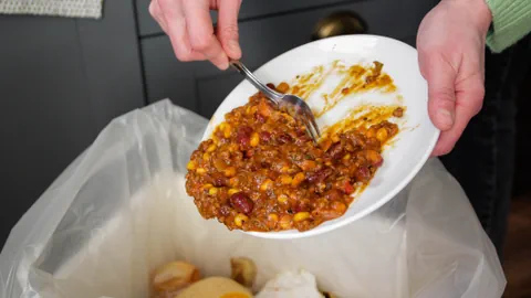 Eine Person lehrt Essen auf einem Teller in einen Abfalleimer.