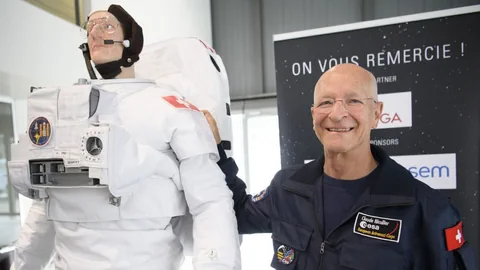 Rechts ist eine Astronautenpuppe zu sehen, daneben steht Claude Nicollier, ein älterer Mann mit Glatze.