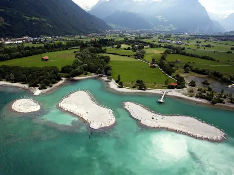 Luftaufnahme von drei Inseln im türkisfarbenen Fluss mit Bergen im Hintergrund.