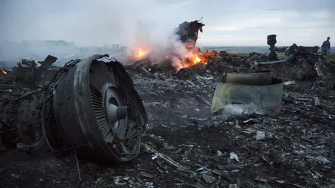 Eine Turbine im Vordergrund, dahinter brennt es: Flugzeugeinzelteile liegen verstreut auf dem Boden.