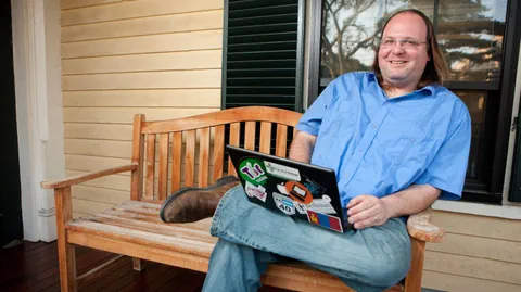 Mann mit schulterlangen Haaren, beginnender Glatze und Brille mit einem Laptop auf dem Schoss