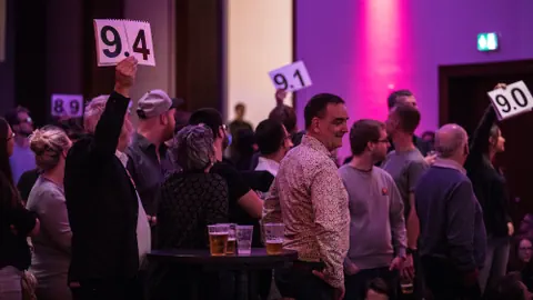 Personen stehen in einem Theatersaal. Einzelne halten Zettel Noten in die Luft.