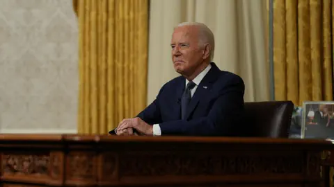 Joe Biden im Anzug sitzt an einem Schreibtisch in einem Raum mit goldenen Vorhängen.