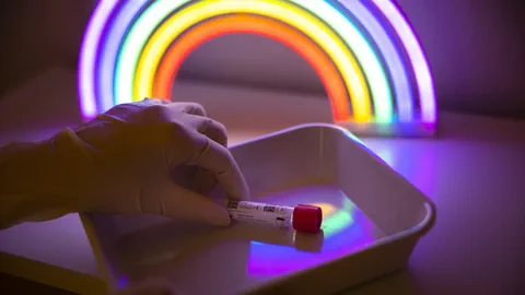 Eine Hand legt eine Ampulle in einen Teller; dahinter ist ein grell leuchtender LED-Regenbogen zu sehen