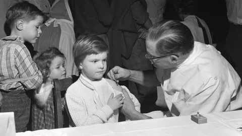 Arzt impft ein Kind mit einer Spritze in den Oberarm. Weitere Kinder warten in einer Schlange.