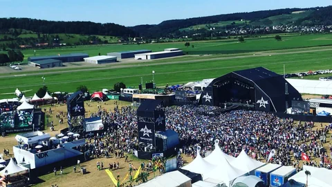 Luftaufnahme eines grossen Musikfestivals mit Bühne, Menschenmenge und Zelten.