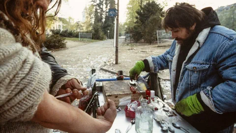 Zwei Männer stehen um einen improvisierten Tisch, einer setzt sich grade eine Spritze in den Arm.