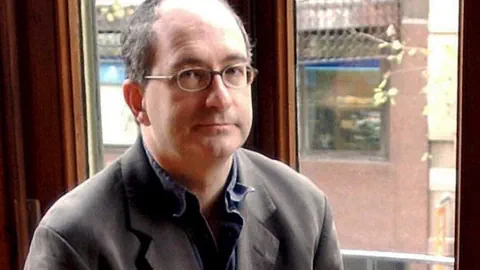 Der britische Autor John Lanchester sitzt in einem dunkelgrauen Anzug auf einem Fenstersims.