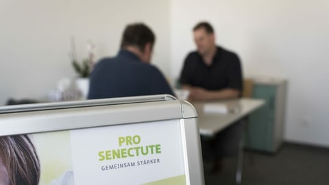 Zwei Personen in einem Gespräch. Im Vordergrund eine Tafel mit dem Logo von Pro Senectute.