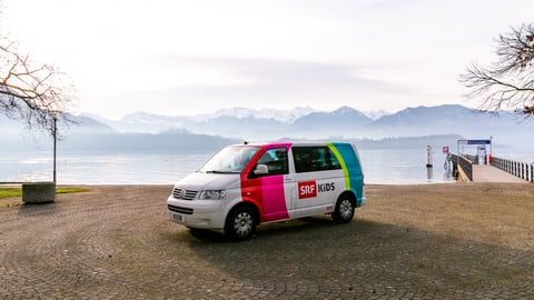 Der SRF Kids Bus steht vor einem See – im Hintergrund sieht man eine Berglandschaft.