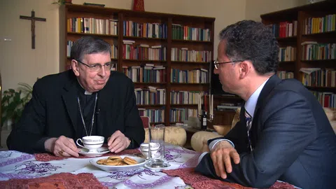 Ein katholischer Würden träger und ein Mann im Anzug sitzem am Tisch und trinken Kaffee. 