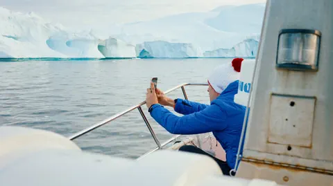 Person in blauer Jacke fotografiert Eisberge von einem Boot aus.