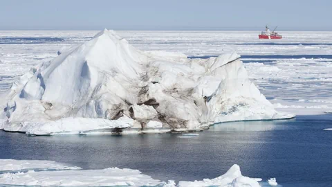 Ein Eisberg liegt in der Arktis auf dem Meer. Dahinter sieht man einen Frrachter. 