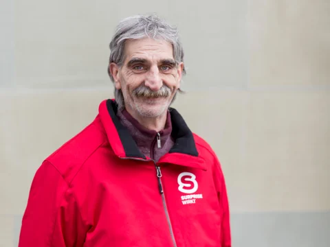 Der Mann Roger Meier in einer roten Surprise Jacke. Er hat einen grossen Schnauz und graue Haar und ist sehr gepflegt.