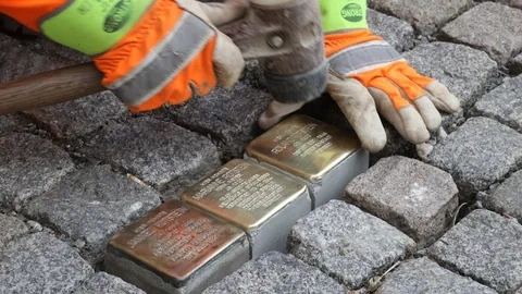 Handwerkerhände, die drei goldene Tafeln im Pflastersteinboden einlassen.