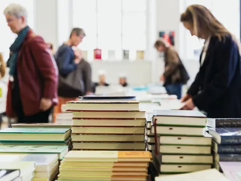 Bücher liegen auf einer Auflage und dahinter schauen sich mehrere Personen die Bücher an.