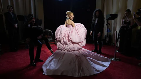 Ariana Grande steht im pinken Kleid auf einem roten Teppich. 