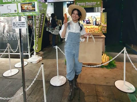 Frau in Latzhose und Strohhut präsentiert Karotte vor dem Stand des Farming Simulators