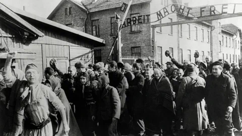 Menschen laufen durch ein Tor und recken zum Teil die Fäuste. Über dem Tor steht: Arbeit macht frei. Es ist Auschwitz.