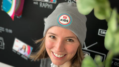 Eine Frau trägt ein graues Beanie mit dem SRF Kids Logo drauf. Sie lächelt in die Kamera. 