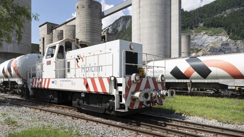 Ein weisser Güterzug mit dem Holcim-Logo steht vor Materialsilos des Holcim Zementwerks.