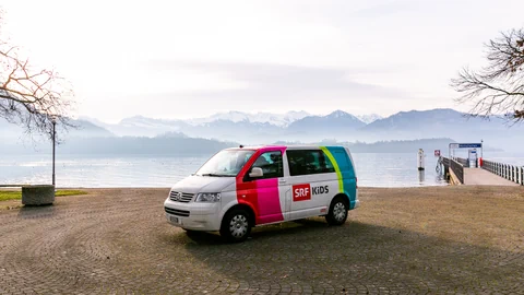 Der SRF Kids Bus steht vor einem See – im Hintergrund sieht man eine Berglandschaft.