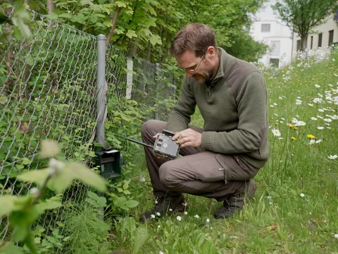 Fabian Kern kauert neben einem Drahtzaun in einer Wiese. Er kontrolliert die Wildkamera, die am Zaun befestigt ist.