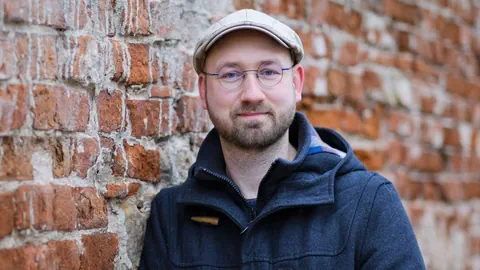 Mann mit Brille und Mütze vor einer Backsteinmauer.