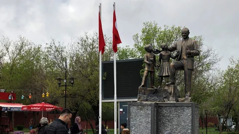 Statue eines Mannes und zwei Kindern auf einem Steinsockel, Passanten, zwei Fahnen und Bäume im Hintergrund