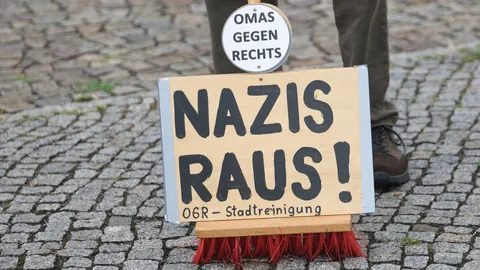 Schild 'Nazis raus!' auf Besen, Kopf mit Schild 'Omas gegen Rechts'.