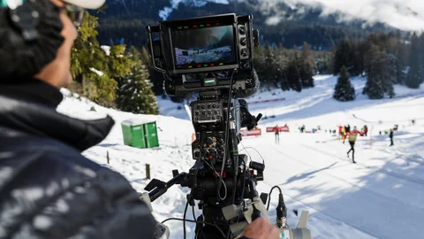 Eine Person steuert eine Kamera, die einen Langlauf-Wettkampf im Hintergrund filmt.