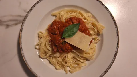 Ein weisser Teller mit Nudeln und roter Tomatensauce. Darauf liegt ein Basilikumblatt und ein Stück Parmesan. 