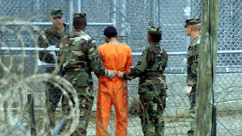 Gefangener in orangefarbener Kleidung von Soldaten begleitet in umzäuntem Bereich.