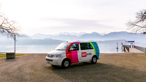 Der SRF Kids Bus vor einem See mit Bergen im Hintergrund.