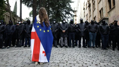 Person mit EU- und georgischer Flagge steht vor einer Gruppe uniformierter Menschen.